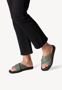 Sandales vertes à brides croisées avec une texture mate douce, semelles noires et un design ouvert, associées à un jean noir effiloché.