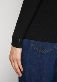 Haut noir côtelé à manches longues avec un poignet ajusté et une petite étiquette de marque, porté avec un jean en denim présentant un design classique de poche arrière.