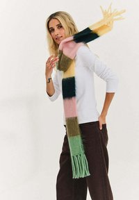 Blondynka w białej koszuli z długim rękawem, ciemnych spodniach i wielokolorowym, paskowanym szaliku z frędzlami, pozująca na jednolitym tle.