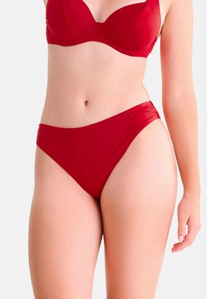 Femme portant un soutien-gorge rouge à armatures et une culotte taille haute assortie, debout devant un fond blanc uni.