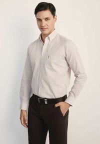 Koszula z długim rękawem zapinana na guziki w jasnym be żowym kolorze w małą kratkę, z klasycznym kołnierzykiem i jedną kieszenią na piersi, zestawiona z ciemnobrązowymi spodniami.