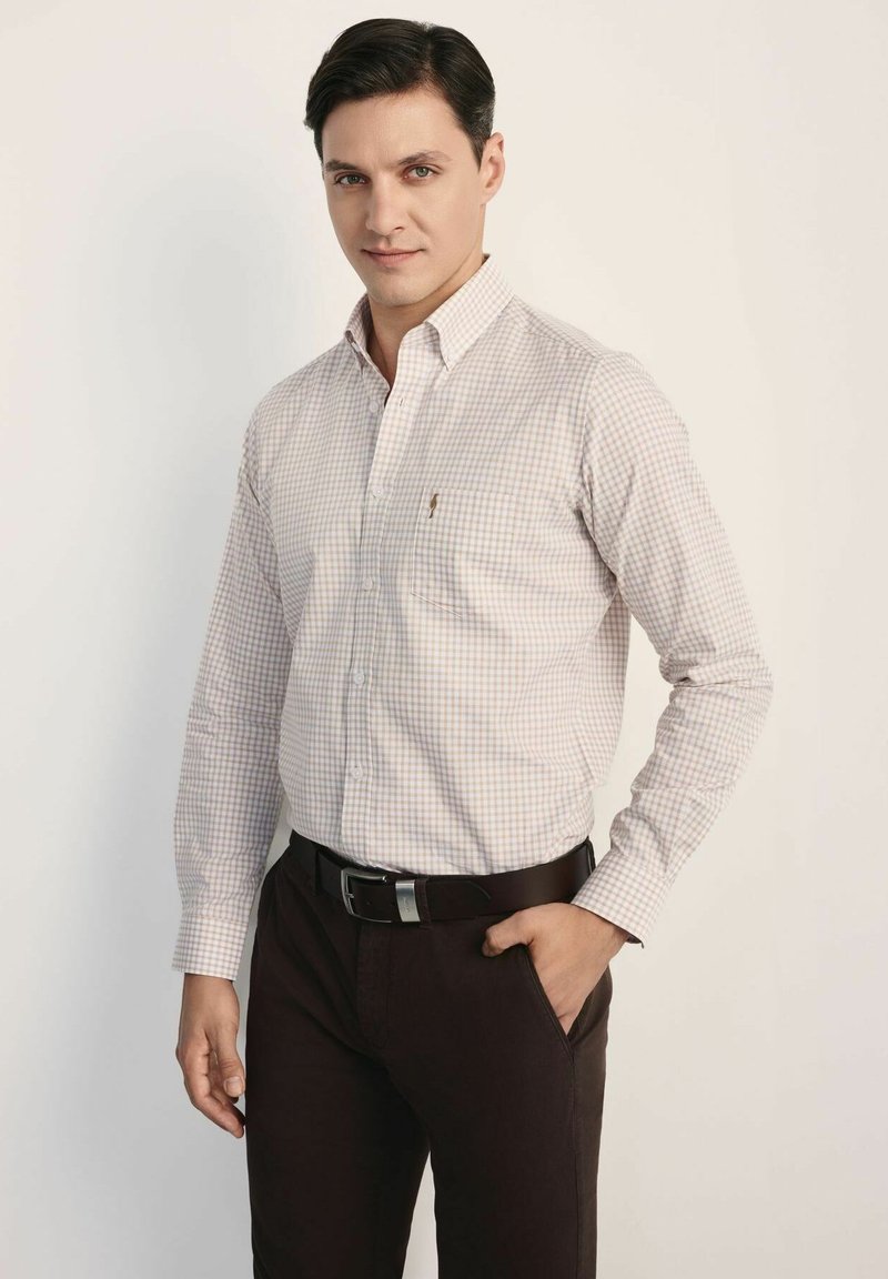 Koszula z długim rękawem zapinana na guziki w jasnym beżowym kolorze w małą kratkę, z klasycznym kołnierzykiem i jedną kieszenią na piersi, zestawiona z ciemnobrązowymi spodniami.