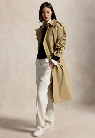 Femeie stă cu mâinile în buzunare purtând un trench bej, pulover negru, cămașă albă, pantaloni albi și adidași albi pe un fundal neutru.
