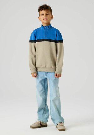 Ragazzo in piedi con le mani rilassate, indossa un maglione con zip blu e beige, jeans azzurri e scarpe da ginnastica beige su uno sfondo neutro.