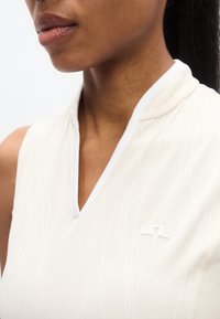 Ανοιχτό μπεζ γιλέκο χωρίς μανίκια με ψηλό γιακά και λευκές λεπτομέρειες. Διαθέτει διακριτό υφασμένο μοτίβο και μικρό λευκό λογότυπο στο στήθος.