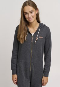 Grå zip-up jumpsuit med hette, med en gyllen glidelås, snorer og et lite logo-patch. Myk tekstur og tettsittende design.