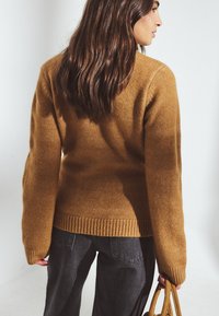 Horčicový pletený sveter s nadmernými rukávmi a rebrovaným lemom, skombinovaný so širokými tmavošedými nohavicami a béžovou kabelkou.