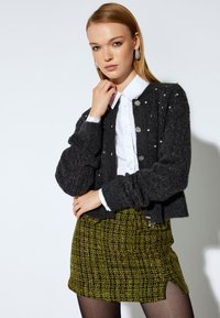 Cardigan gris de punto con un patrón texturizado y embellishments, combinado con una camisa blanca de cuello y una falda mini a cuadros verde.