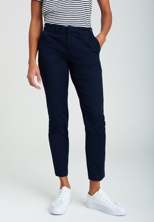 Personne portant un pantalon court bleu marine ajusté, des baskets blanches et un haut à manches courtes rayé noir et blanc, debout avec une main dans la poche.