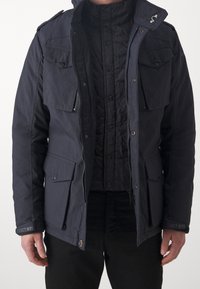 Granatowa kurtka z wieloma kieszeniami, zapięciem na suwak i pikowaną podszewką. Posiada kaptur oraz mankiety z oznaczeniem marki.