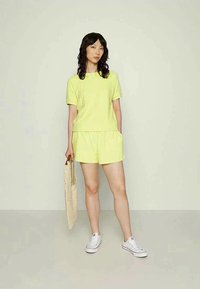 T-shirt de algodão verde limão com mangas curtas e calções combinando, apresentando um corte relaxado. Saco de tecido e ténis brancos completam o look.