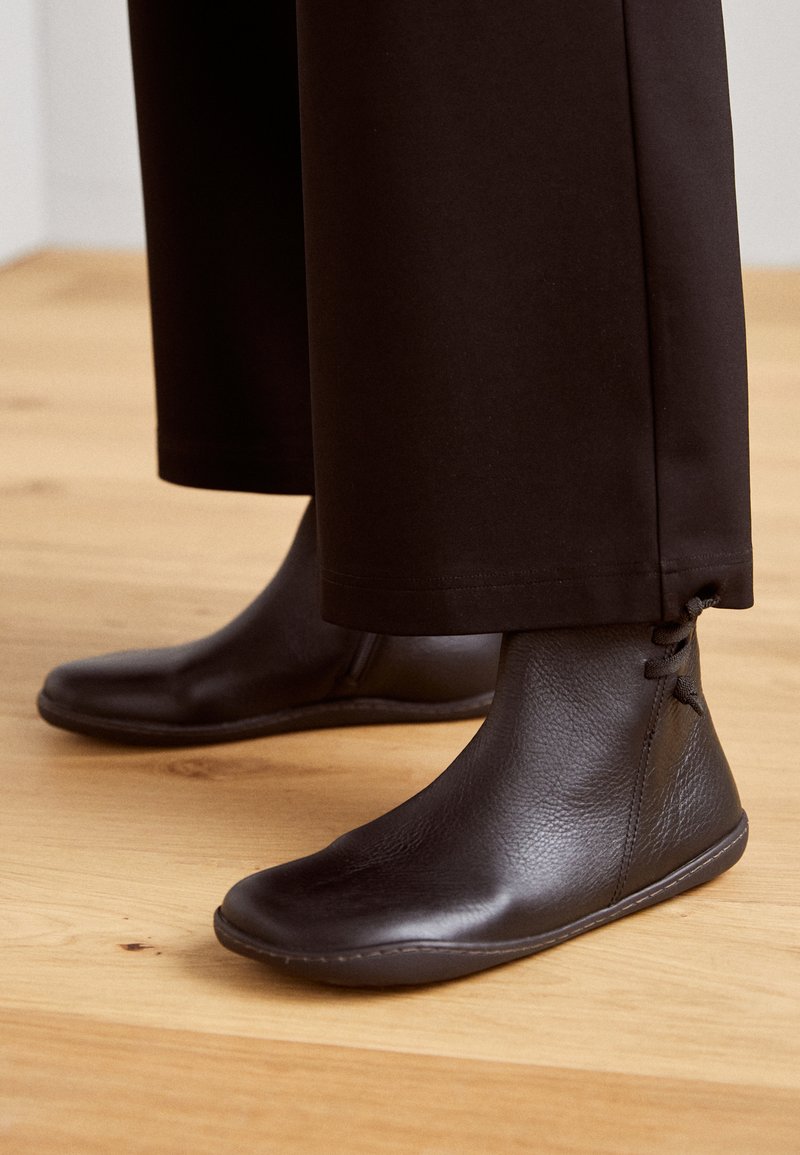 Black leather ankle boots with a textured finish, flat soles, and lace detail at the back, paired with wide black pants on a wooden floor.