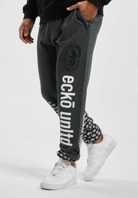 Grå sweatpants med en afslappet pasform, hvide logo-tryk langs siden og mønstrede detaljer ved manchetterne. Båret med hvide sneakers.