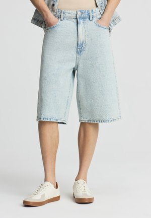 Uomo che indossa pantaloncini di jeans azzurri lavati fino al ginocchio con tasche, sneakers bianche con suole marroni e una maglietta beige parzialmente visibile.