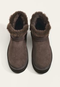 Smešasti smeđi čizmi do gležnja od suede materijala s umetkom od umjetnog krzna na rubu, zaobljenog oblika i ojačanim đonom. Glatka tekstura s vidljivim detaljima šavova.