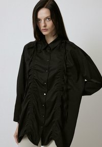 Black shirt with a relaxed fit, featuring a button-up front, ruffled texture down the center, and long sleeves with subtle gathers.