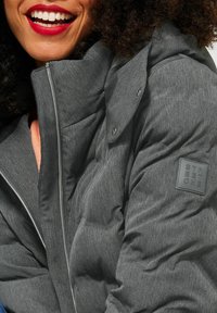 Grå vadderad jacka med ett strukturerat tyg, dragkedjestängning, stor krage och logotypmärke med texten "STREET ONE" på ärmen.