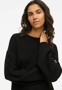Sweater preto com textura canelada, mangas largas e decote redondo. O tecido parece macio e quente, projetado para conforto.