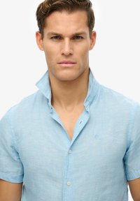 Jasnoniebieska koszula lniana z krótkim rękawem i kołnierzykiem zapinanym na guziki, z delikatną teksturą i małym wyszywanym logo na piersi.