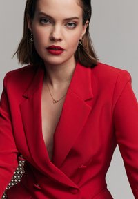 Czerwony żakiet z głębokim dekoltem, z metalowymi guzikami w kolorze srebrnym i gładką fakturą. Złoty naszyjnik w kształcie serca oraz kolczyki w kształcie kół uzupełniają look.
