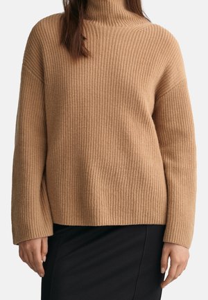Kvinde iført en beige ribstrikket rullekravebluse med lange ærmer og et sort skørt, stående foran en ensfarvet baggrund.