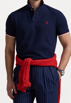 Man draagt marineblauw poloshirt met rood logo, rode trui om de taille gebonden, blauwe broek met fijne strepen en polshorloge aan linkerarm.