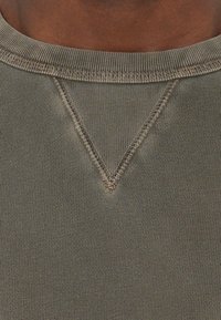 Gros plan sur un pull vert à col rond avec un détail de couture en forme de V distinctif sous le col sur une peau brun foncé.