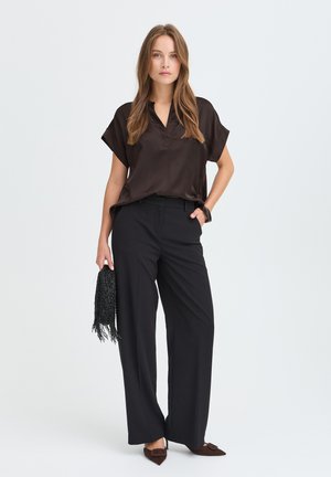 Femme portant un chemisier marron foncé et un pantalon noir à jambes larges, tenant un sac à main noir à franges, debout devant un fond uni.
