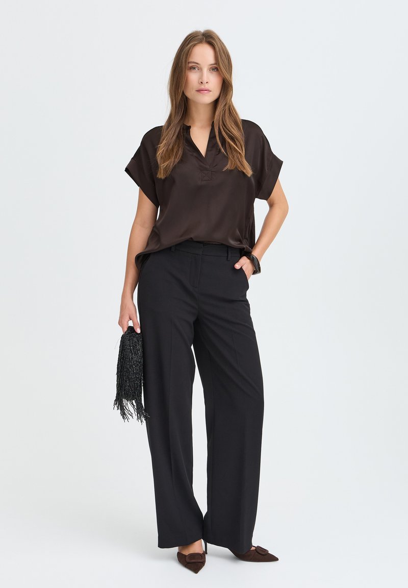 Femme portant un chemisier marron foncé et un pantalon noir à jambes larges, tenant un sac à main noir à franges, debout devant un fond uni.