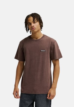 Młody mężczyzna z zaplecionymi włosami, ubrany w zwykły brązowy t-shirt z krótkim rękawem i ciemne dżinsy na białym tle.
