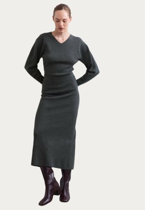 Kvinde iført en mørkegrå langærmet sweater og matchende midi nederdel kombineret med mørkebrune støvler med hæl, stående med hænderne på ryggen.