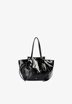 Bolso tipo tote de cuero negro y brillante con dos asas y un pequeño emblema dorado de calavera en el frente sobre un fondo blanco.