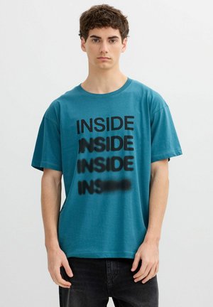 Joven con camiseta verde azulado con la palabra "INSIDE" impresa cuatro veces, cada línea se difumina progresivamente hacia abajo.