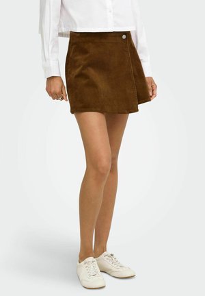 Femme portant une mini-jupe en velours côtelé marron avec un bouton latéral, une chemise blanche à manches longues et des chaussures blanches à lacets, debout sur un fond blanc.