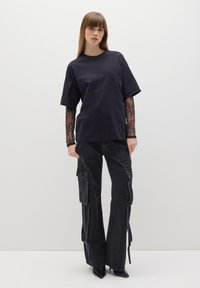 Camiseta negra de cuello redondo combinada con pantalones cargo negros que cuentan con bolsillos, cremalleras y tacones negros puntiagudos. Las mangas largas de encaje transparente son visibles.