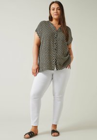 Blusa de manga corta con estampado en blanco y negro, diseño de cuello en V, combinada con pantalones ajustados blancos y sandalias negras planas, de pie sobre un fondo claro.