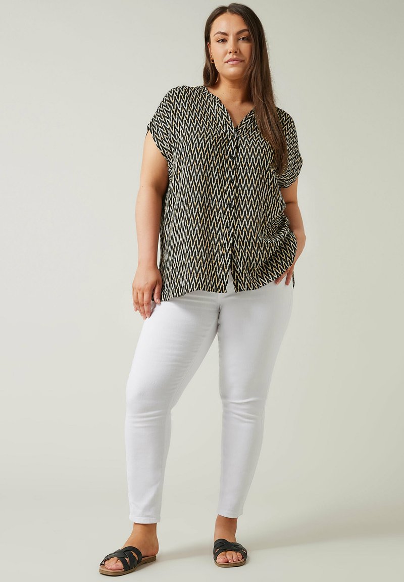 Blusa de manga corta con estampado en blanco y negro, diseño de cuello en V, combinada con pantalones ajustados blancos y sandalias negras planas, de pie sobre un fondo claro.