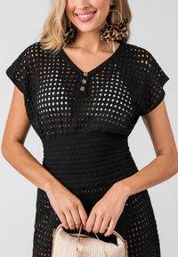 Abito all'uncinetto nero con scollatura a V, caratterizzato da un design allentato e perforato e tre bottoni in legno. La modella tiene in mano una borsa intrecciata.