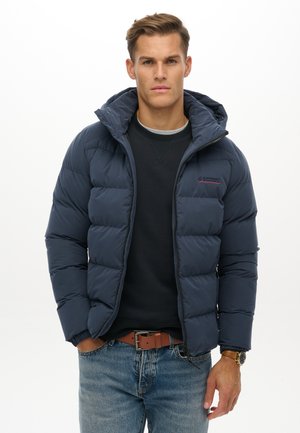 Junger Mann trägt eine marineblaue Steppjacke über einem schwarzen Sweatshirt und blauen Jeans, mit braunem Gürtel und Armbanduhr, steht vor weißem Hintergrund.