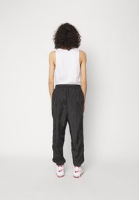 Schwarze Sporthose mit elastischem Bund, kombiniert mit einem weißen Tanktop. Das Modell trägt weiße Sneaker mit roten Akzenten. Ansicht von hinten.