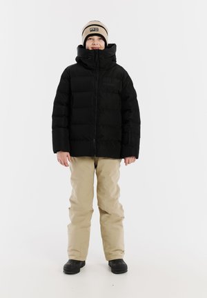 Schwarze Puffjacke mit hohem Kragen, beigen Hosen und schwarzen wasserdichten Stiefeln. Die Jacke verfügt über einen Reißverschluss auf der Vorderseite und seitliche Taschen.