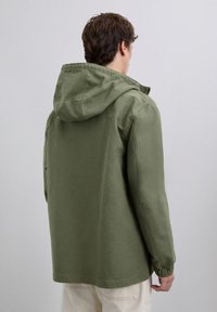 Homme portant une veste à capuche olive et un pantalon beige clair, tourné vers le dos devant un fond gris clair uni.