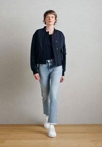 Tamsiai mėlyna bombonešio striukė su trumpintu dizainu, priekyje užsegama mygtukais ir su kišenėmis, derinama prie šviesiai mėlynų, dėvėtų stiliaus džinsų ir baltų sportinių batelių.