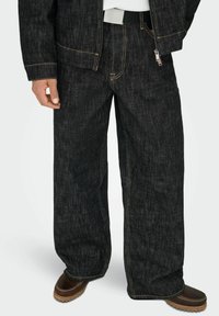 Melnas platajiem džinsi ar kontrastējošām dzeltens šuvēm, aprīkoti ar rāvējslēdzēja aizdari un jostu. Kombinēti ar brūnām slip-on kurpēm.