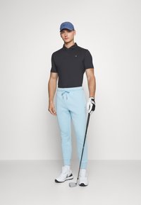 Camisa polo preta, calças de moletom azul claro, sapatilhas brancas e um boné azul. Segurando um taco de golfe, de pé contra um fundo simples.