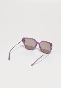 Ochelari de soare purpurii cu un cadru masiv, geometric. Accente din plastic texturat și metal subliniază templele și brațele. Lentile de culoare maro.