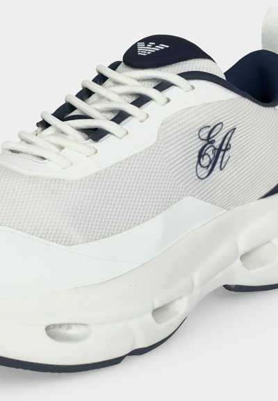 Baskets blanches et bleu marine en tissu texturé, lacets et logo stylisé « EA » sur le côté, avec une semelle épaisse et sculptée dotée de trous de ventilation.