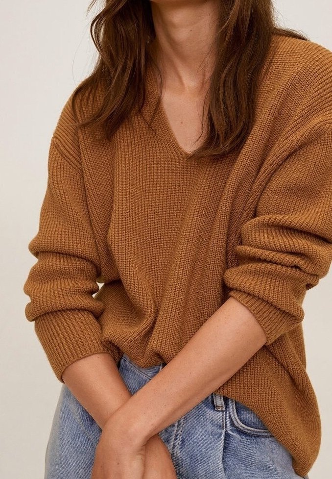 Femme portant un pull en maille côtelée marron avec manches retroussées et un jean bleu clair, posant les bras croisés devant.