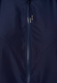 Närbild av en marinblå jacka med dragkedja, med dubbla metalldragkedjehandtag och synlig diagonalsöm på den släta tyget.