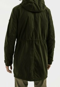 Hombre con una chaqueta parka con capucha de color verde oscuro y cintura ajustada, visto desde atrás sobre un fondo claro y liso.
